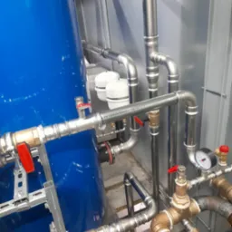 Złożona instalacja wodna z rur stalowych i mosiężnych, z zaworami z czerwonymi uchwytami, manometrem i niebieskim zbiornikiem ciśnieniowym. Widok z bliska, oświetlenie techniczne.