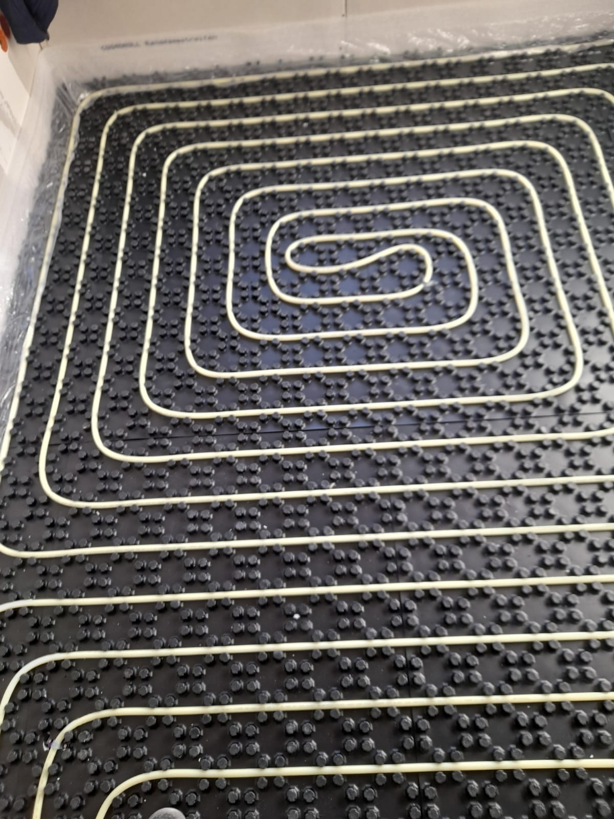 Ułożona instalacja ogrzewania podłogowego na czarnej macie z wypustkami, widok z góry. Białe rurki tworzą spiralny wzór na powierzchni.