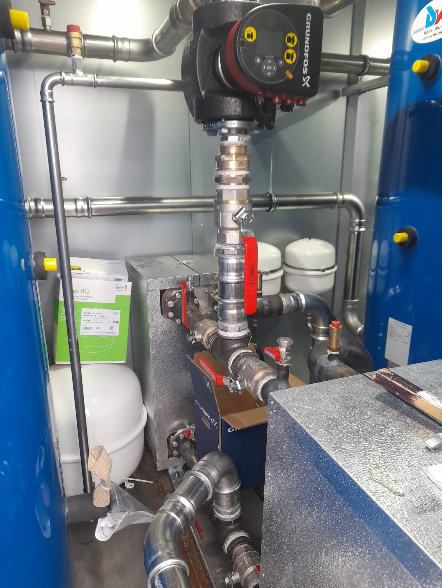 Węzeł ciepłowniczy z pompą obiegową Grundfos, zbiornikami wyrównawczymi i orurowaniem. Widoczne zawory, manometry i elementy instalacji w pomieszczeniu technicznym.