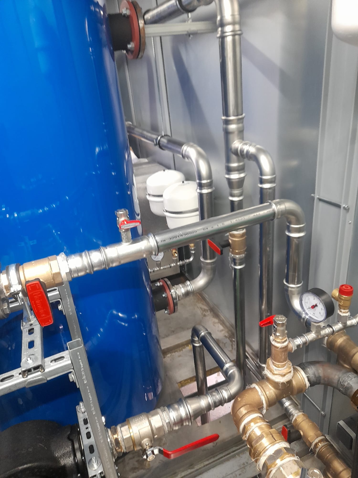 Złożona instalacja wodna z rur stalowych i mosiężnych, z zaworami z czerwonymi uchwytami, manometrem i niebieskim zbiornikiem ciśnieniowym. Widok z bliska, oświetlenie techniczne.