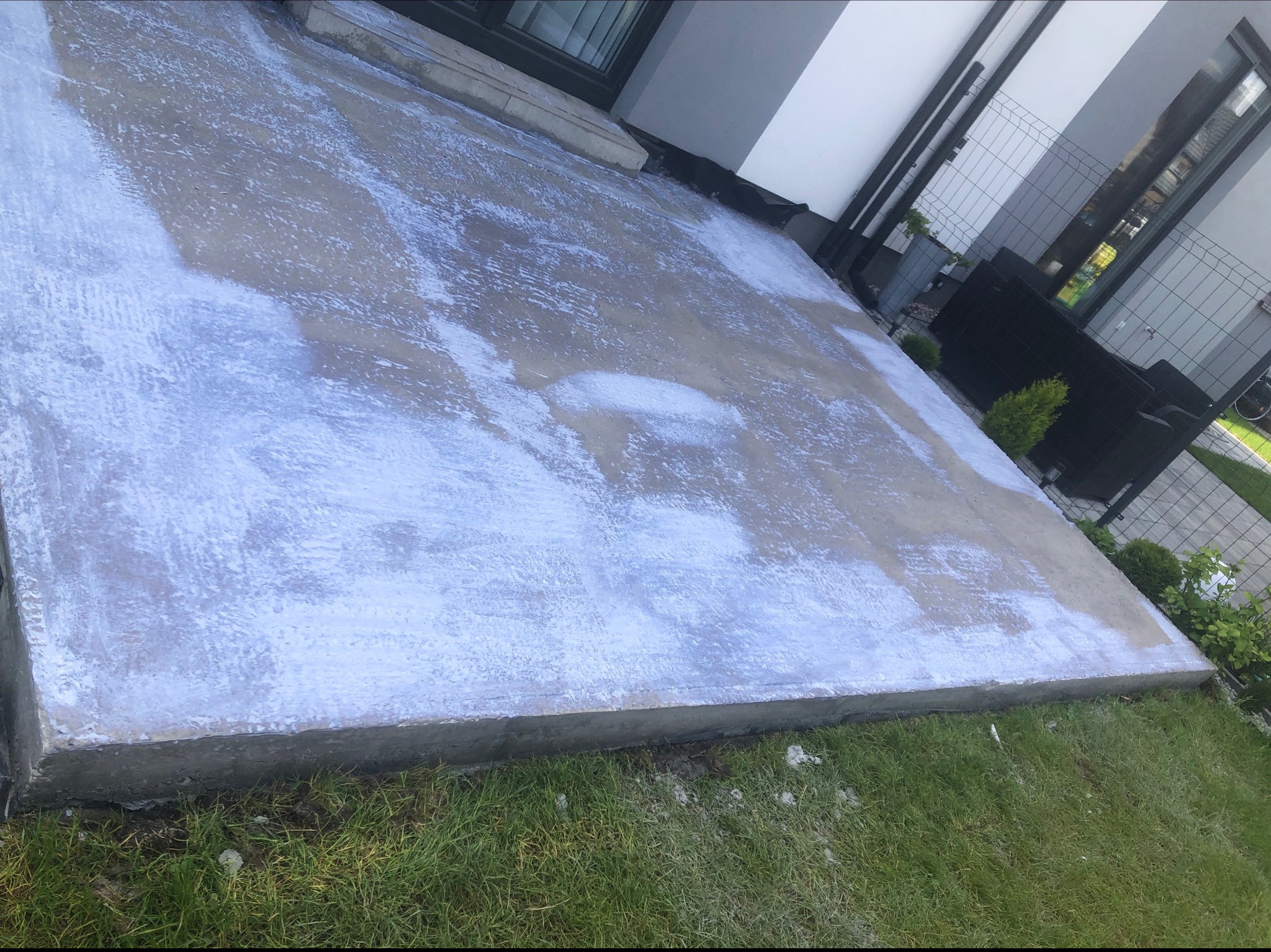 Świeżo wykonany taras betonowy z widocznymi śladami białej farby gruntującej, otoczony zielonym trawnikiem i elementami architektury ogrodowej.