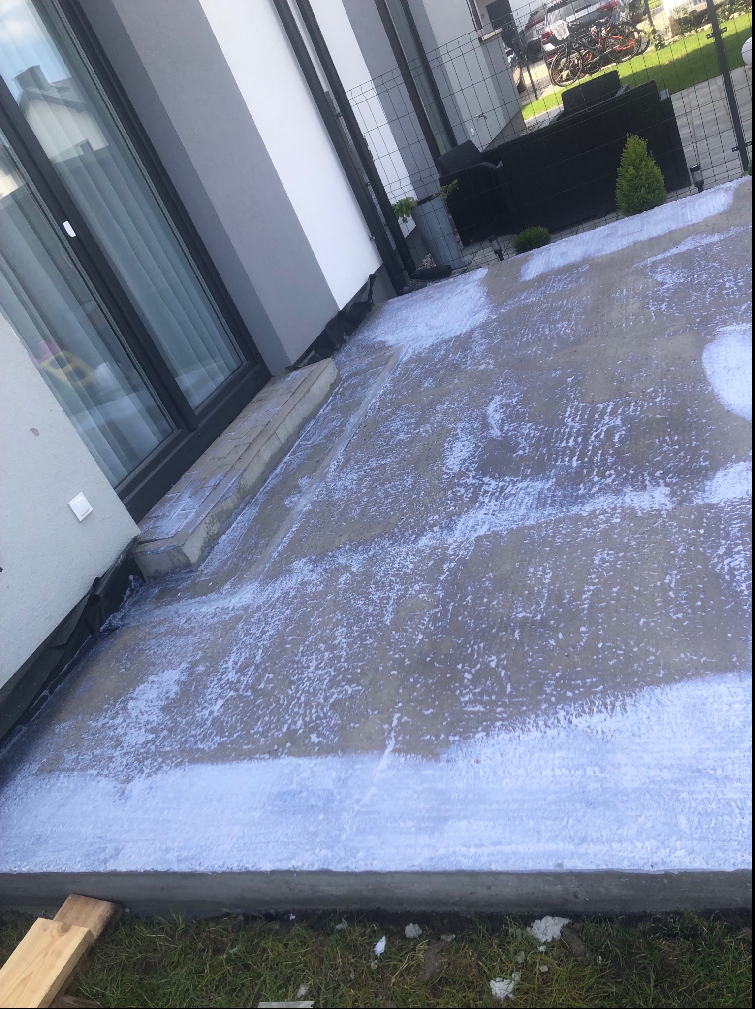 Świeża, wilgotna wylewka betonowa na tarasie przylegającym do domu jednorodzinnego, z widocznymi śladami po zacieraniu. Na drugim planie meble ogrodowe.