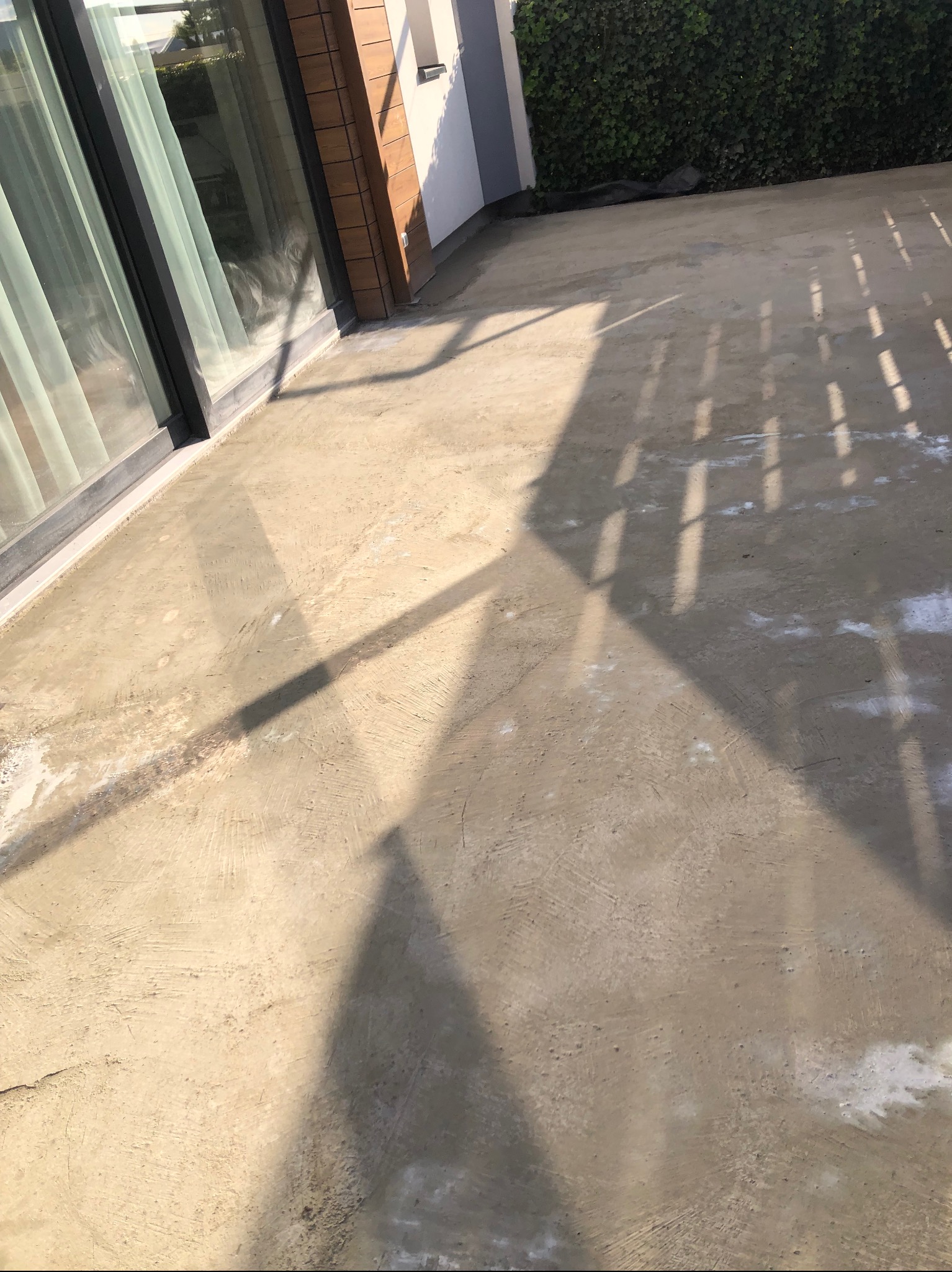 Świeża, jasna wylewka na tarasie przylegającym do budynku z dużym oknem. Na powierzchni widoczne cienie rzucane przez konstrukcję zadaszenia.
