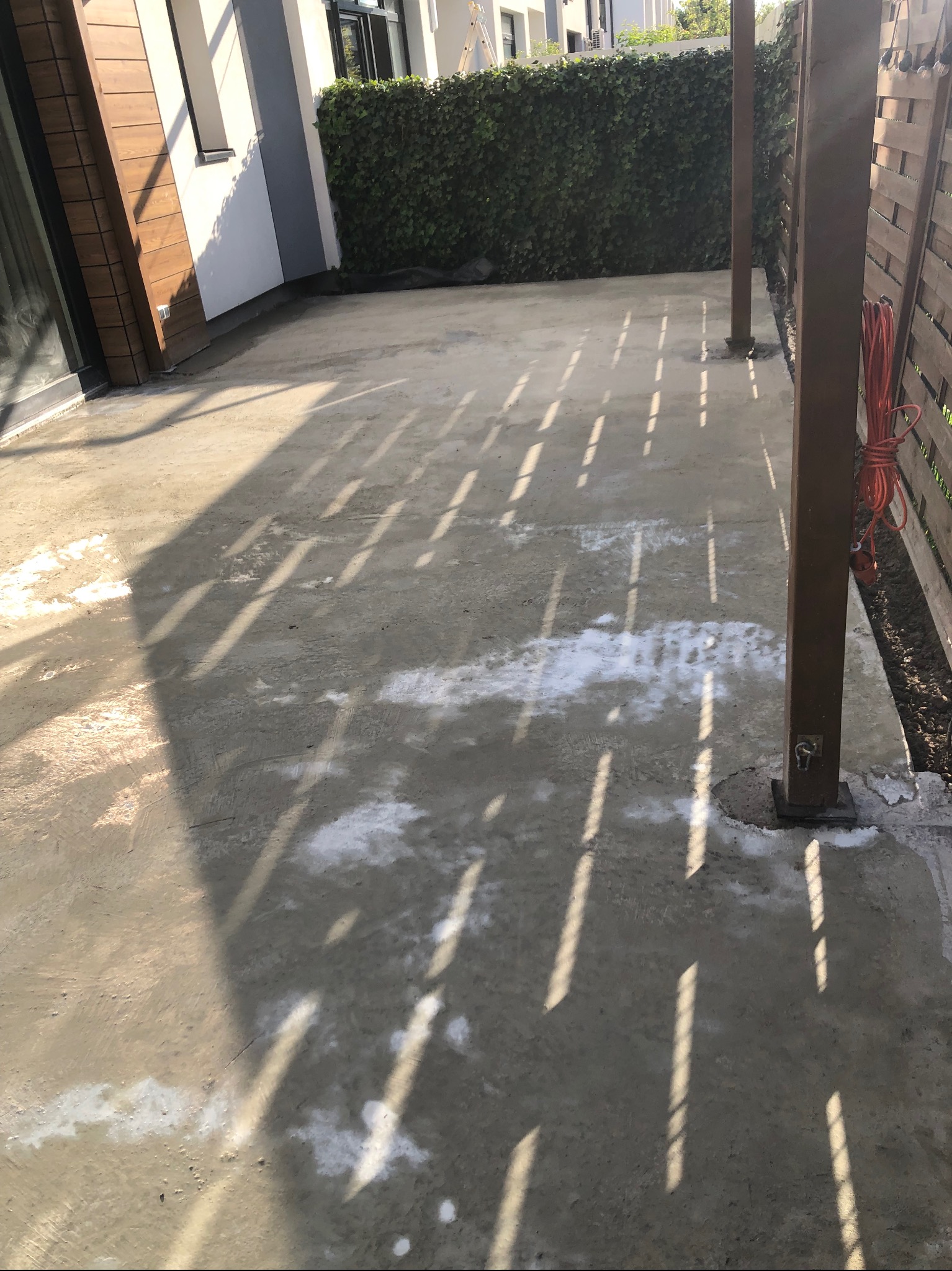 Świeża, szara wylewka na zewnątrz z jasnymi smugami światła słonecznego, drewniane słupy i zielony żywopłot w tle.