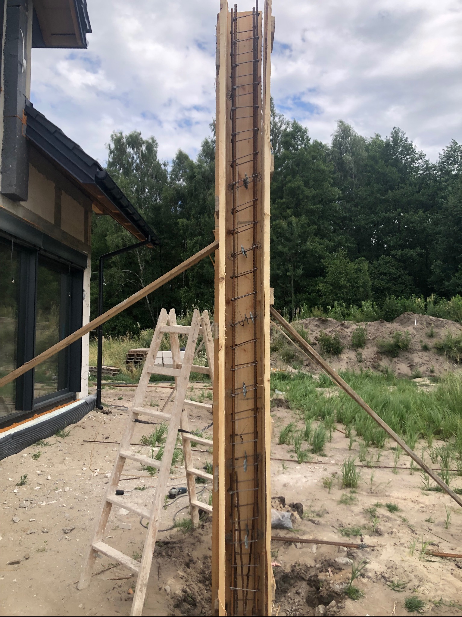 Szczegółowe ujęcie zbrojenia kolumny w drewnianym szalunku przy budynku w budowie, drabina oparta o konstrukcję, zieleń w tle. Widok na etap prac.
