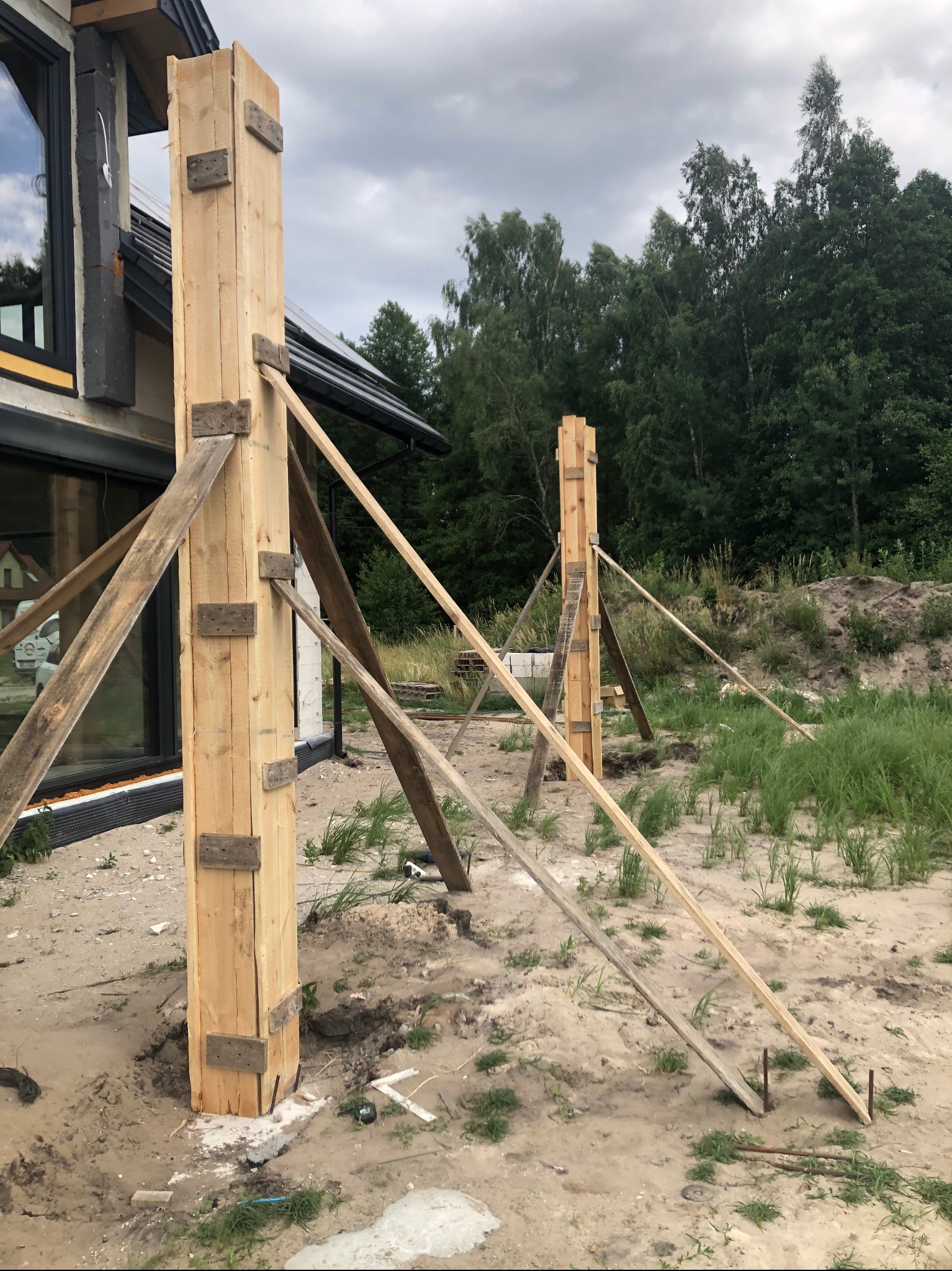 Drewniane szalunki kolumnowe, podparte skośnymi belkami, na tle domu z dużymi oknami i zieleni. Etap przygotowawczy do wylewania betonu.