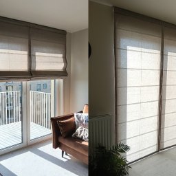 Szymon Makowski Rolele - Beżowe rolety rzymskie z tkaniny na oknie balkonowym, z widokiem na budynek. Obok sofa z poduszkami i roślina doniczkowa. Dwie rolety zamontowane obok siebie.