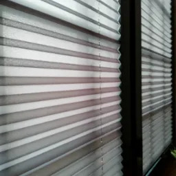 Szare plisy okienne zamontowane w ramie okiennej, widoczne z bliska, z delikatnym efektem światłocienia na plisowanej strukturze materiału.