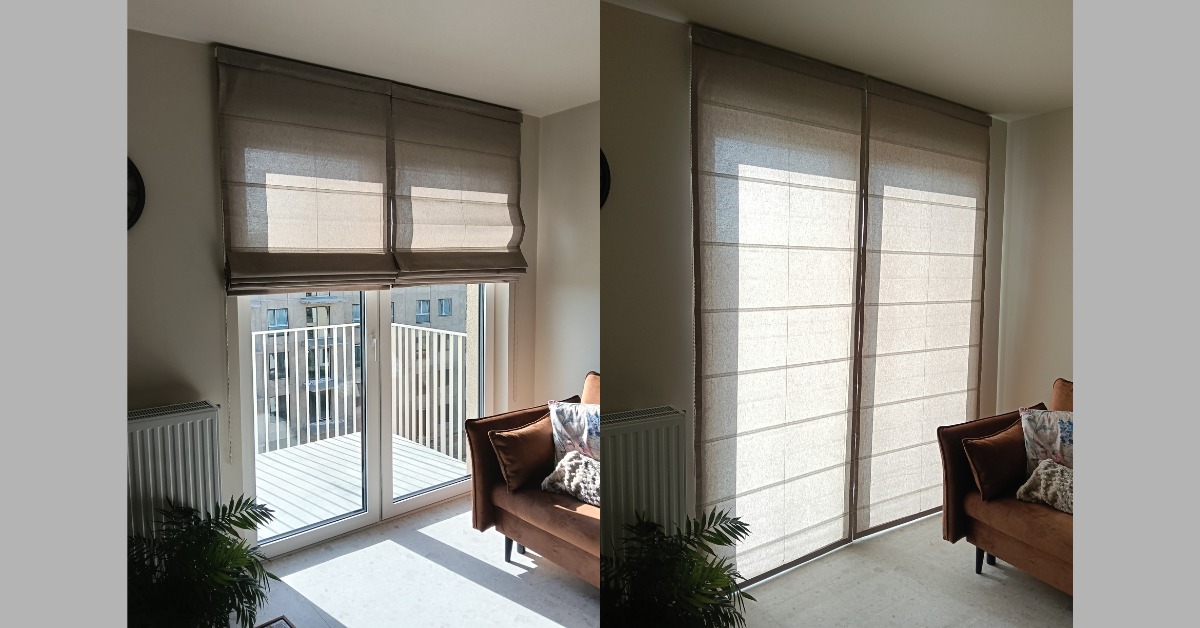 Beżowe rolety rzymskie z tkaniny na oknie balkonowym, z widokiem na budynek. Obok sofa z poduszkami i roślina doniczkowa. Dwie rolety zamontowane obok siebie.