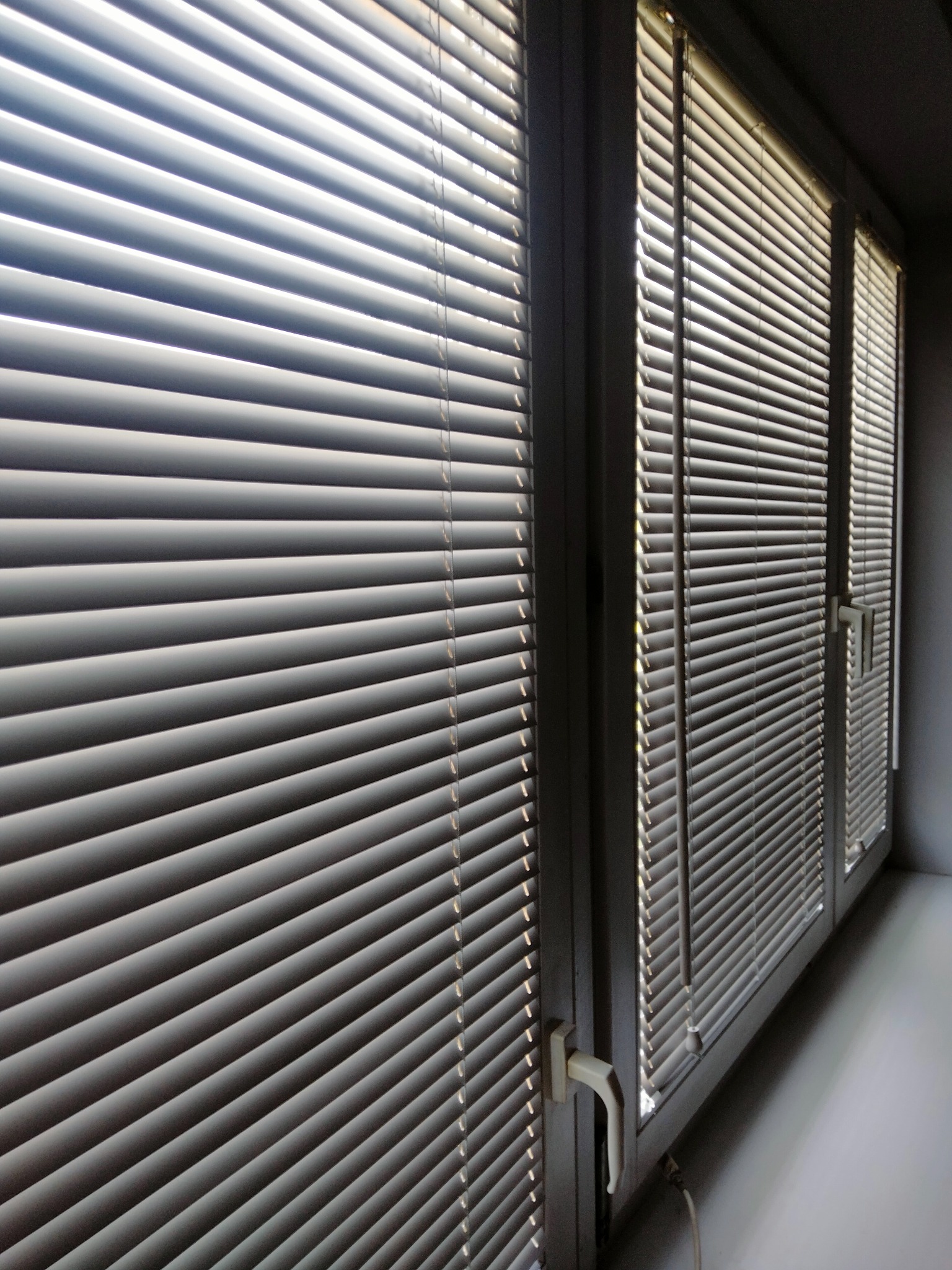 Trzy okna z aluminiowymi żaluzjami w kolorze jasnoszarym, z widocznymi sznurkami i mechanizmami sterującymi, ustawione pod kątem w jasnym pomieszczeniu.