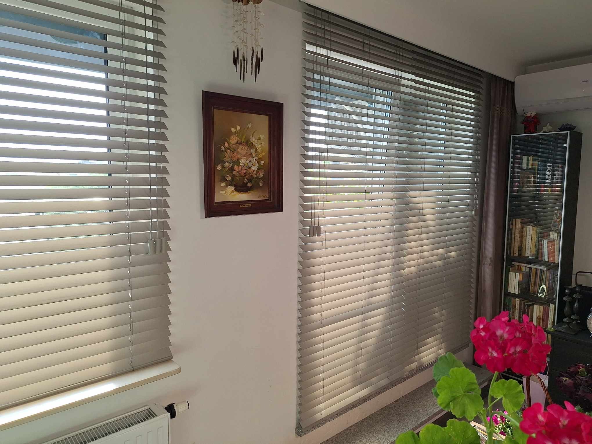 Jasne, poziome żaluzje aluminiowe w oknie, z obrazem kwiatów na ścianie. Oświetlenie słoneczne podkreśla fakturę żaluzji i dodaje ciepła wnętrzu.