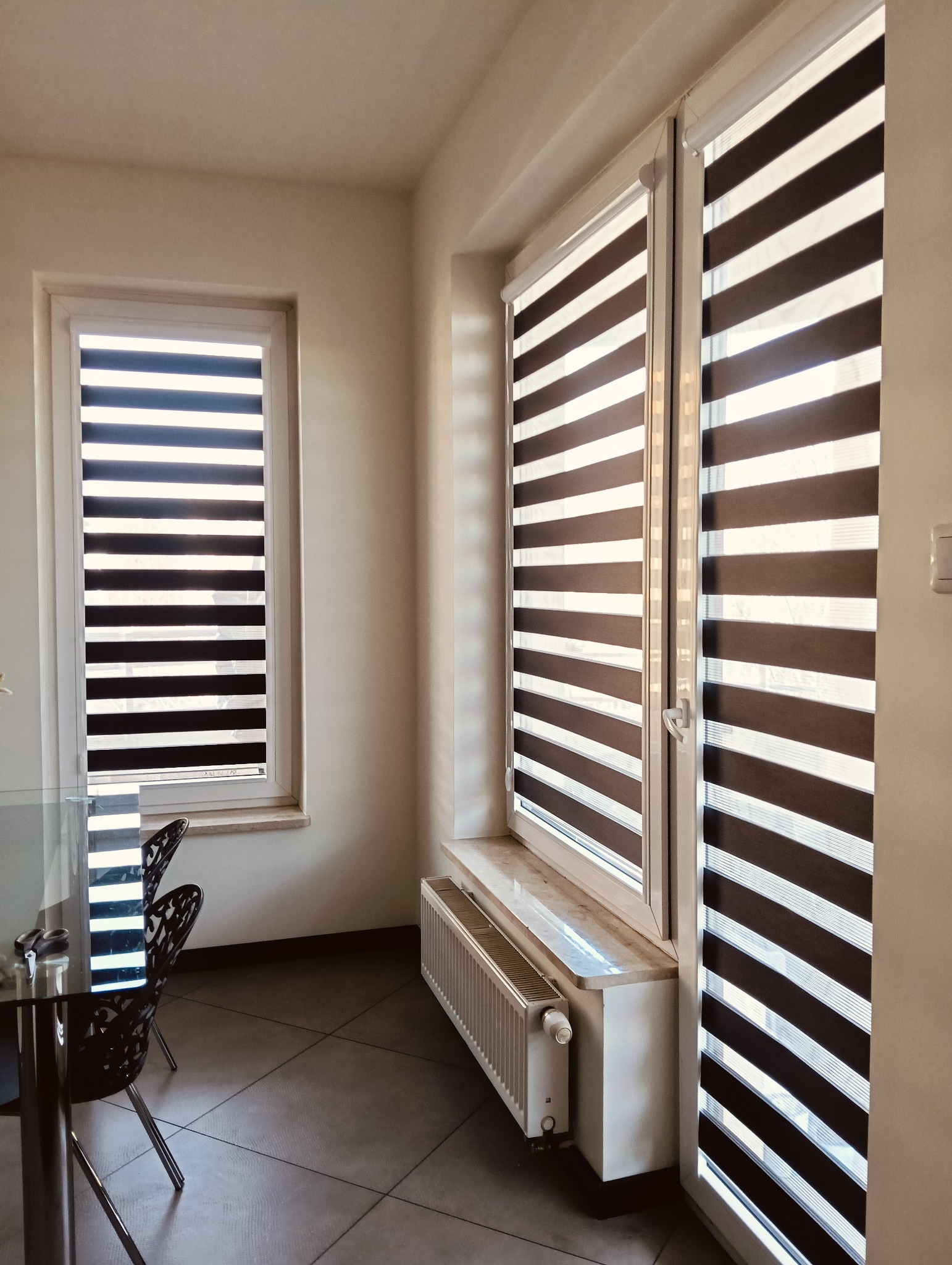 Nowoczesne rolety dzień noc w jasnym wnętrzu. Widoczne dwa okna z zamontowanymi roletami, z charakterystycznymi poziomymi pasami. Na podłodze szare płytki.