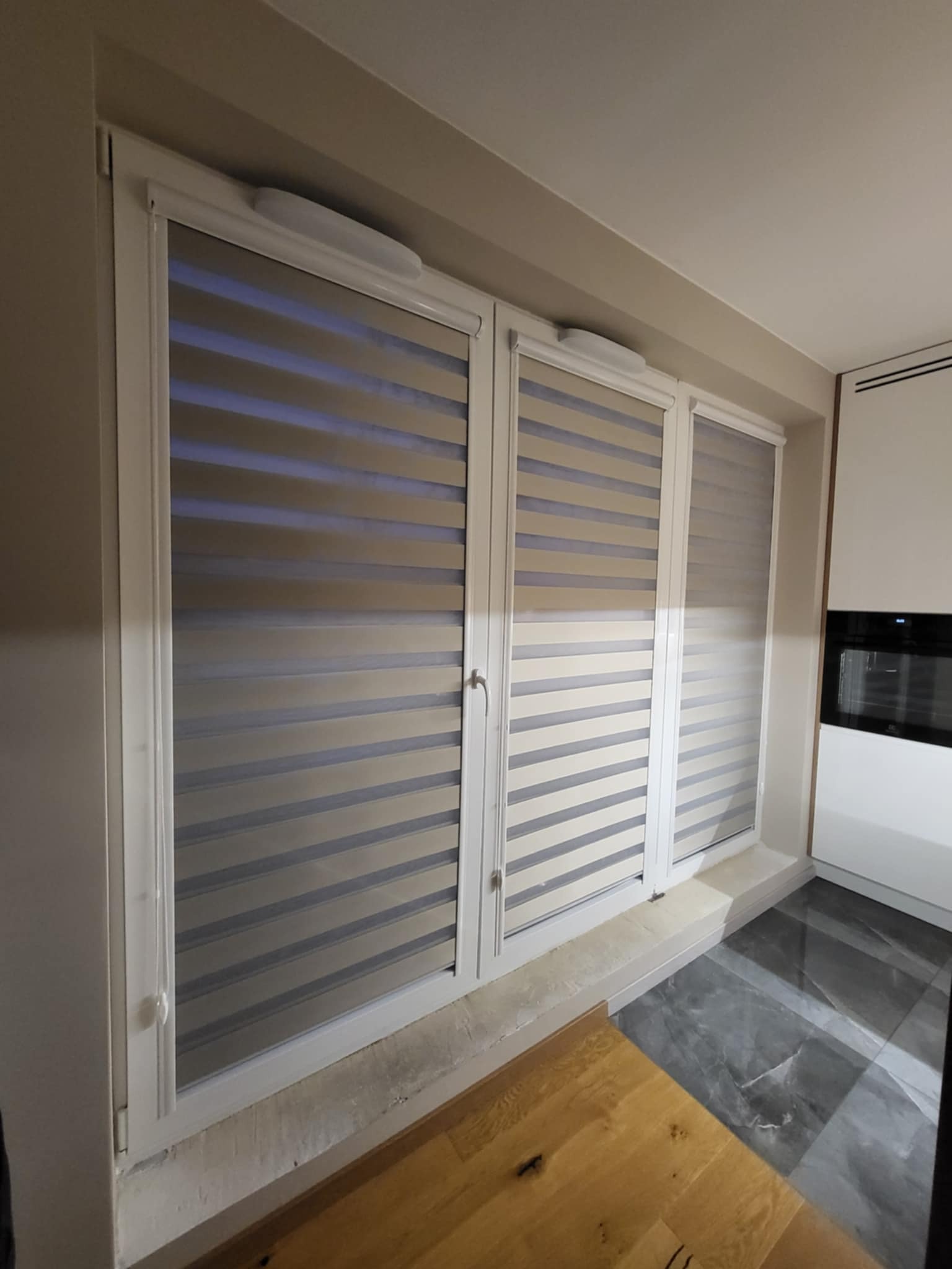 Trzy okna z zamontowanymi szarymi roletami dzień i noc w nowoczesnym wnętrzu. Widoczny fragment podłogi z płytek i parkietu.