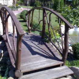 Drewniany mostek ogrodowy z balustradą z gałęzi, prowadzący nad oczkiem wodnym, otoczony zielenią i kamieniami. Prywatne zamówienie.