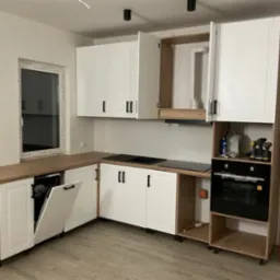 Nowoczesna kuchnia z białymi frontami i drewnianym blatem. Widoczny piekarnik i płyta grzewcza. Część szafek otwarta, ukazująca zawartość. Poziomica oparta o ścianę.