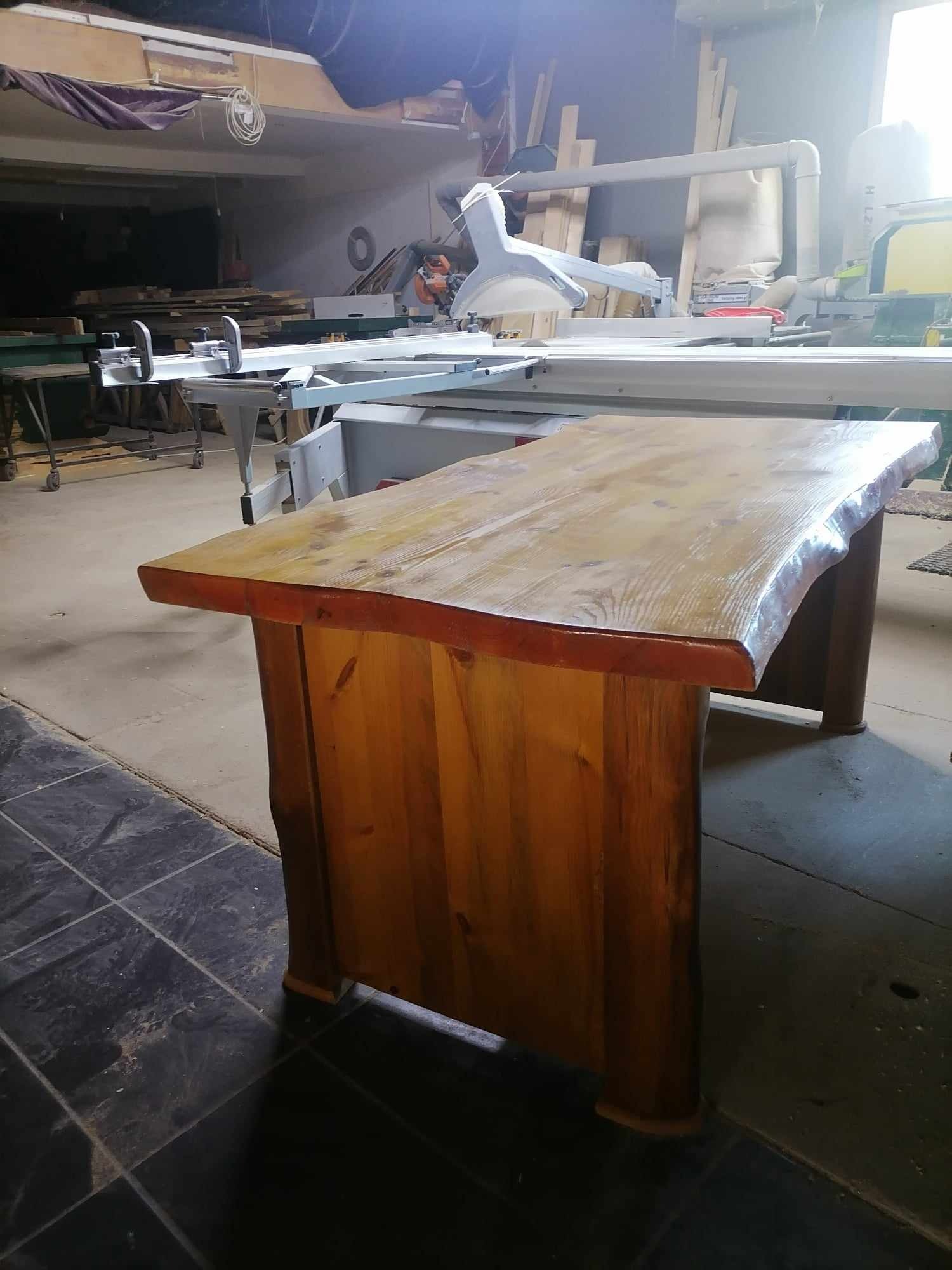 Drewniany stół z blatem o nieregularnych krawędziach, wykonany w warsztacie stolarskim. Widoczne narzędzia i materiały stolarskie w tle.