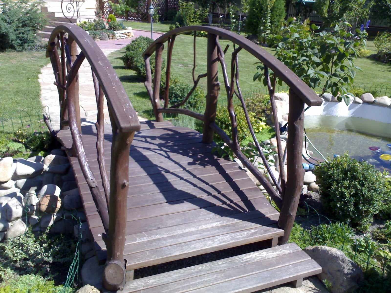 Drewniany mostek ogrodowy z balustradą z gałęzi, prowadzący nad oczkiem wodnym, otoczony zielenią i kamieniami. Prywatne zamówienie.