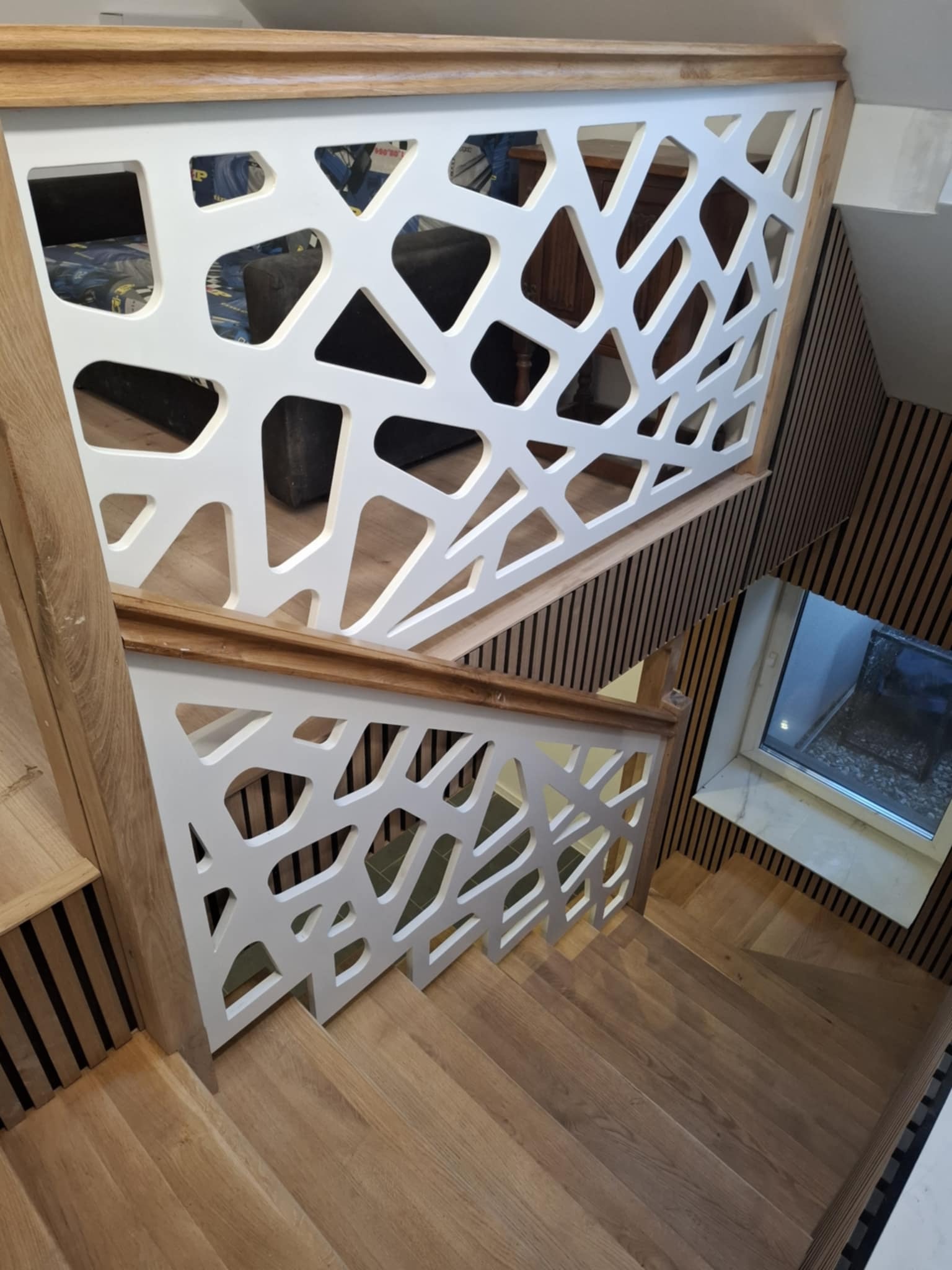 Schody drewniane z ażurową białą balustradą i drewnianymi stopniami. Widok z góry, z elementami dekoracyjnymi na ścianie. Naturalne światło z okna.