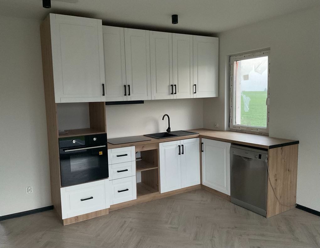 Nowoczesna kuchnia w bieli i drewnie: zabudowa z piekarnikiem, zlewem i zmywarką, jasne wnętrze z oknem na zewnątrz. Parkiet ułożony w jodełkę.