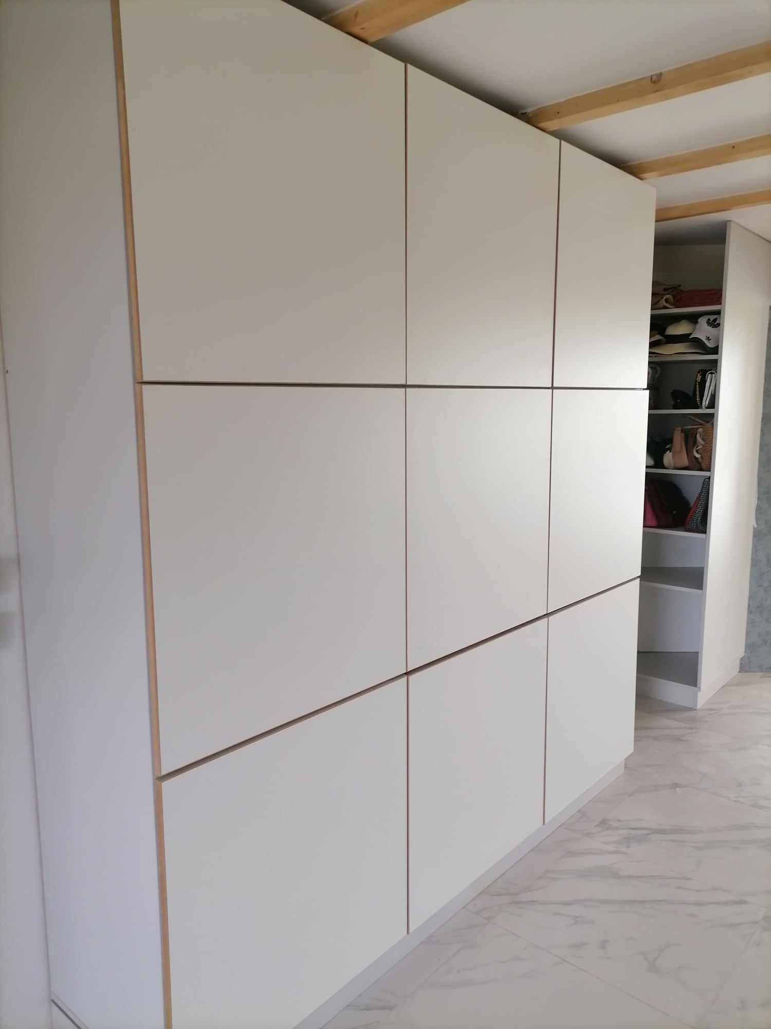 Biała zabudowa meblowa z szafkami, widoczna część garderoby z półkami i akcesoriami. Minimalistyczny design, jasne wnętrze, marmurowe płytki na podłodze.