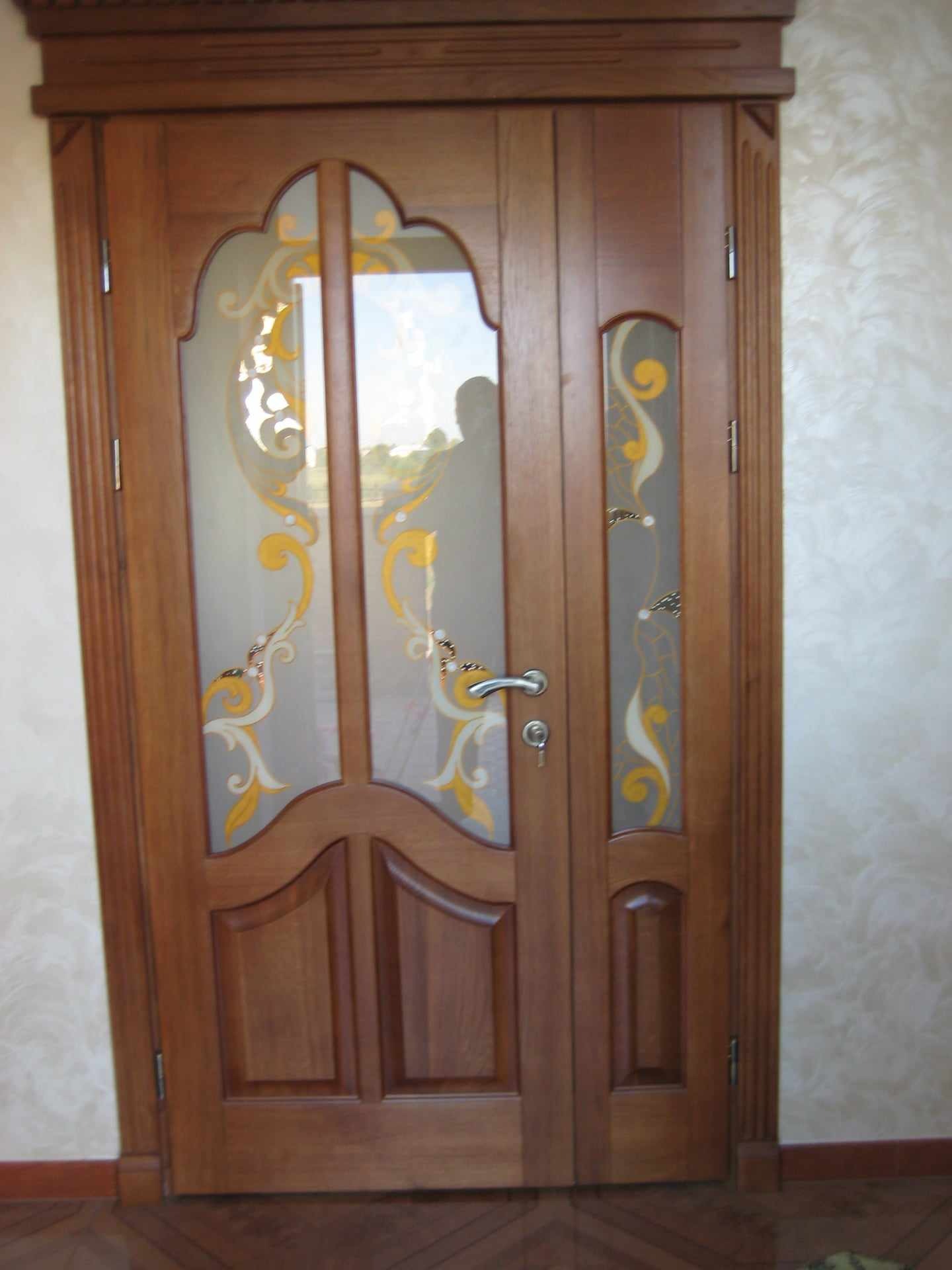 Dwuskrzydłowe, drewniane drzwi z dekoracyjnymi szybami w odcieniach złota i bieli, z metalową klamką i zamkiem, na tle ściany z delikatnym wzorem.