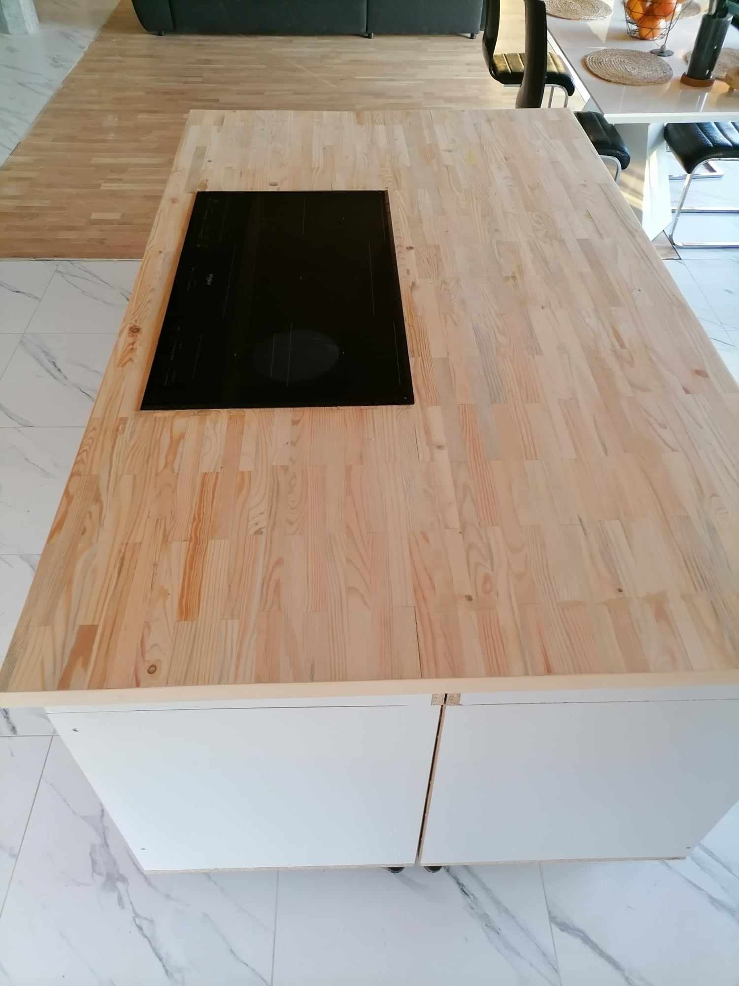 Blat kuchenny z jasnego drewna klejonego, z wbudowaną czarną płytą indukcyjną, na tle parkietu i marmurowej podłogi. Nowoczesna aranżacja kuchni.