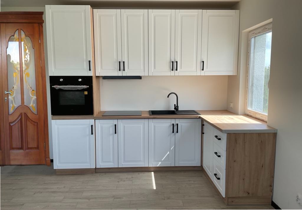 Jasna kuchnia z białymi szafkami i drewnianym blatem, czarny zlew i armatura. Widoczne okno i ozdobne drzwi. Nowoczesny design.