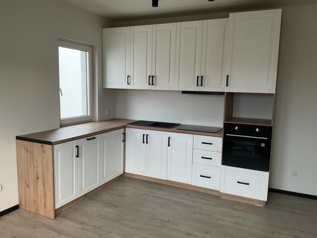 Jasna kuchnia z białymi szafkami i czarnymi uchwytami. Blat drewniany, okno z widokiem na zewnątrz. Nowoczesny minimalizm w neutralnej kolorystyce.