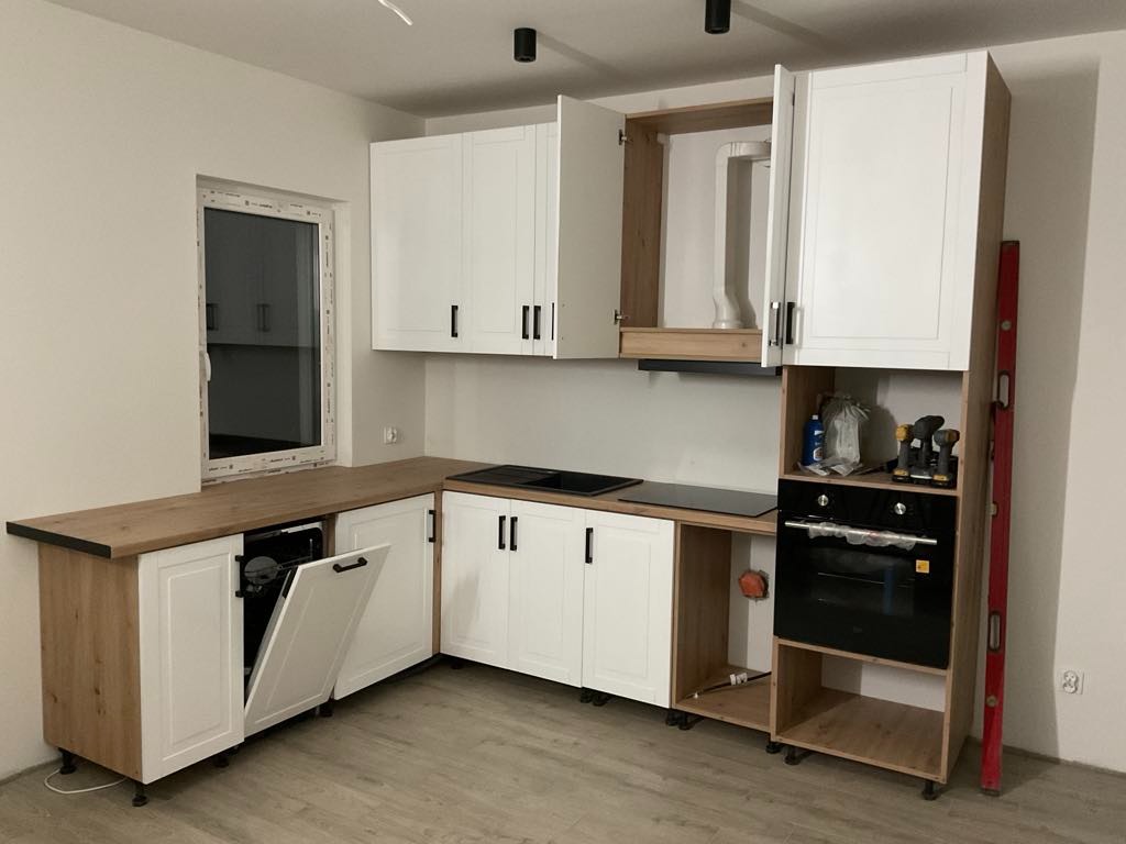 Nowoczesna kuchnia z białymi frontami i drewnianym blatem. Widoczny piekarnik i płyta grzewcza. Część szafek otwarta, ukazująca zawartość. Poziomica oparta o ścianę.