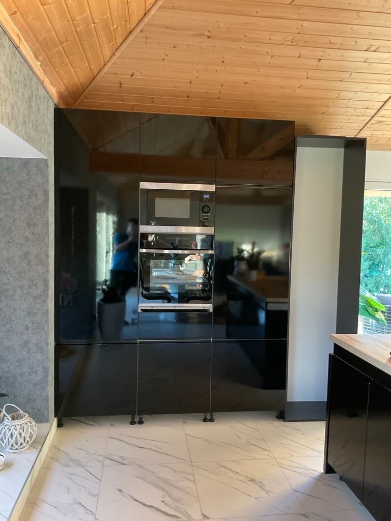 Czarna, lakierowana zabudowa kuchenna z wbudowanym piekarnikiem i mikrofalówką, marmurowa podłoga, drewniany sufit, nowoczesny design, zlecenie prywatne.