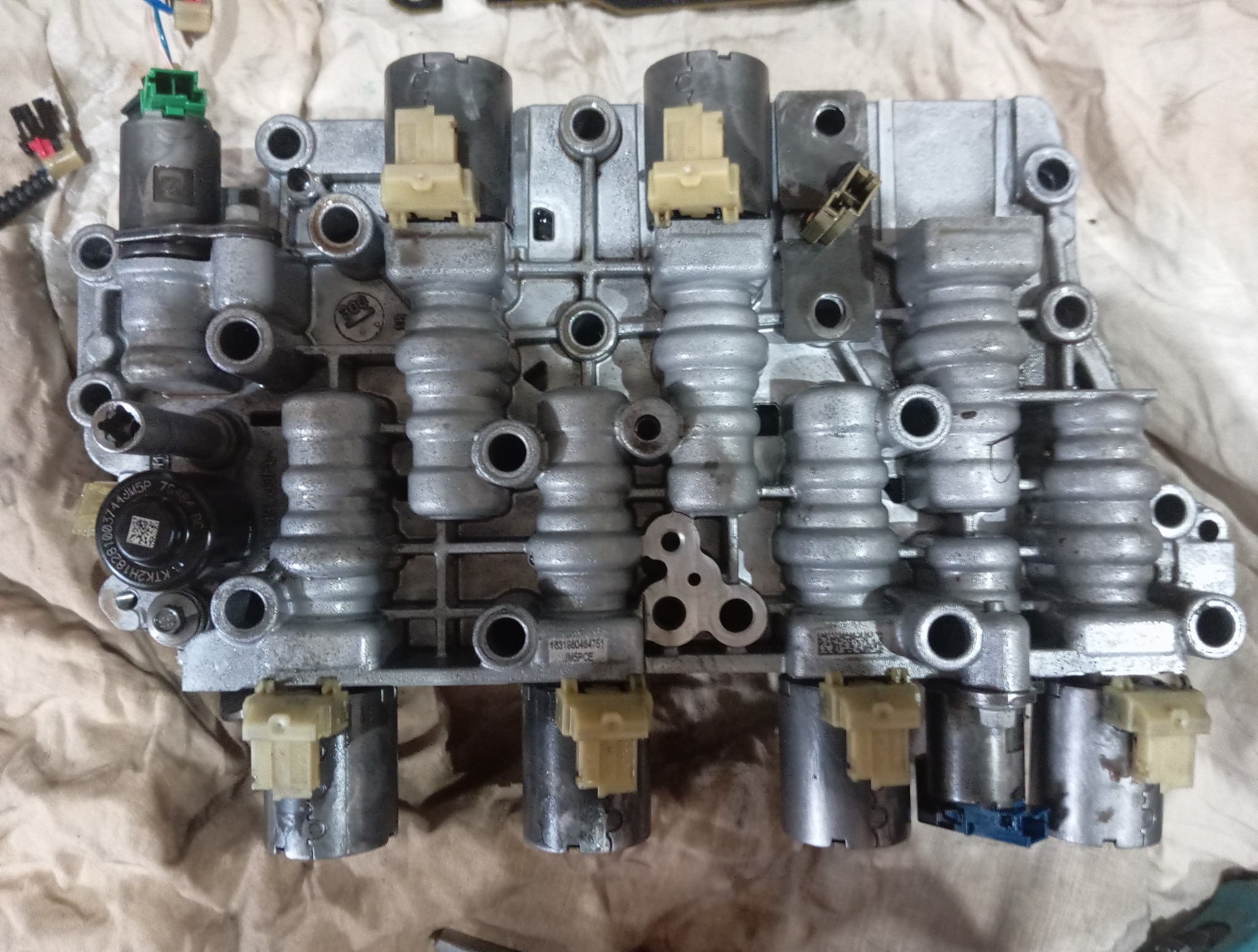 Szara, metalowa płyta sterująca skrzyni biegów z widocznymi solenoidami i złączami elektrycznymi, leżąca na beżowej szmatce. Widoczne numery seryjne na elementach.