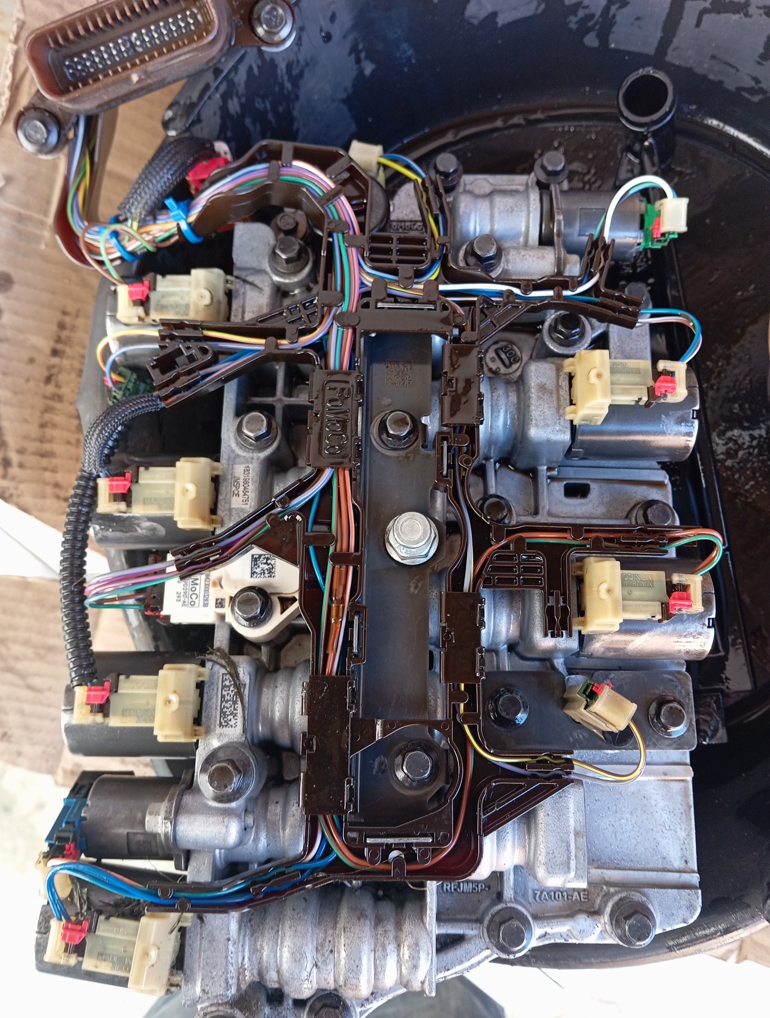 Otwarta skrzynia biegów z widocznymi przewodami i elektroniką w zbliżeniu. Elementy są pokryte olejem. Widoczne oznaczenia FoMoCo i napisy Made in Mexico.