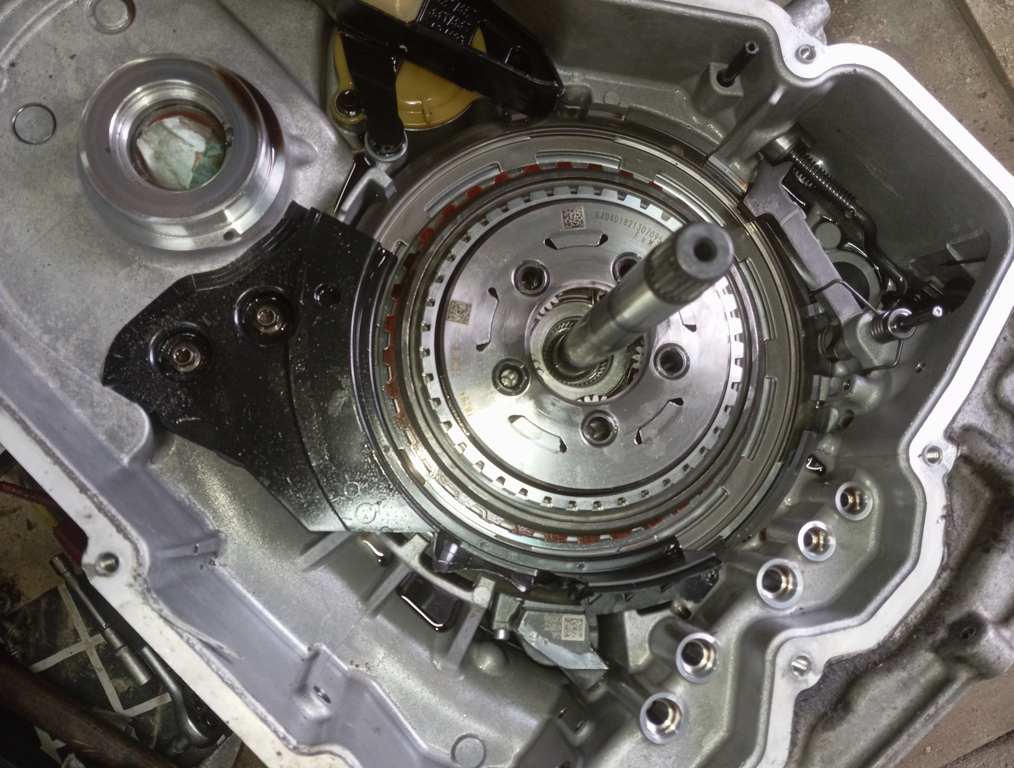 Otwarta skrzynia biegów z widocznymi elementami wewnętrznymi, w tym tarczami i wałem, w srebrnej obudowie. Widoczne ślady oleju i narzędzia w tle.