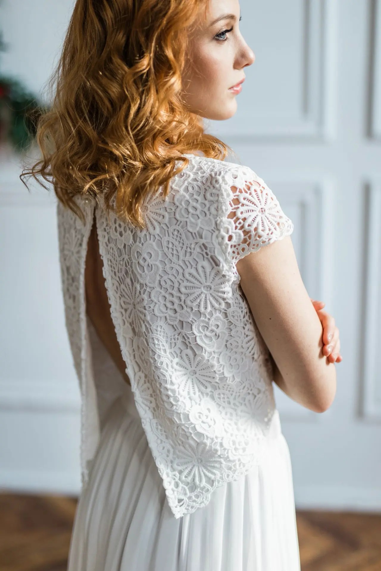 Kobieta z rudymi włosami, ubrana w białą, koronkową suknię z odkrytymi plecami, prezentuje styl rustykalny w jasnym wnętrzu.