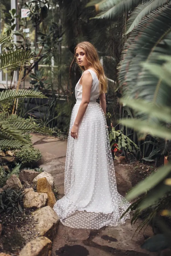 Kobieta w białej sukni ślubnej z tiulowym dołem w kropki, odwrócona w egzotycznym ogrodzie botanicznym. Romantyczna stylizacja, naturalne światło.
