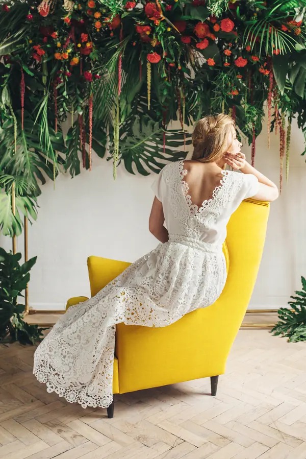 Kobieta w koronkowej sukni ślubnej siedzi na żółtym fotelu, tyłem do kamery. Kwiatowa dekoracja w tle. Suknia z odkrytymi plecami i delikatnym wzorem koronki.
