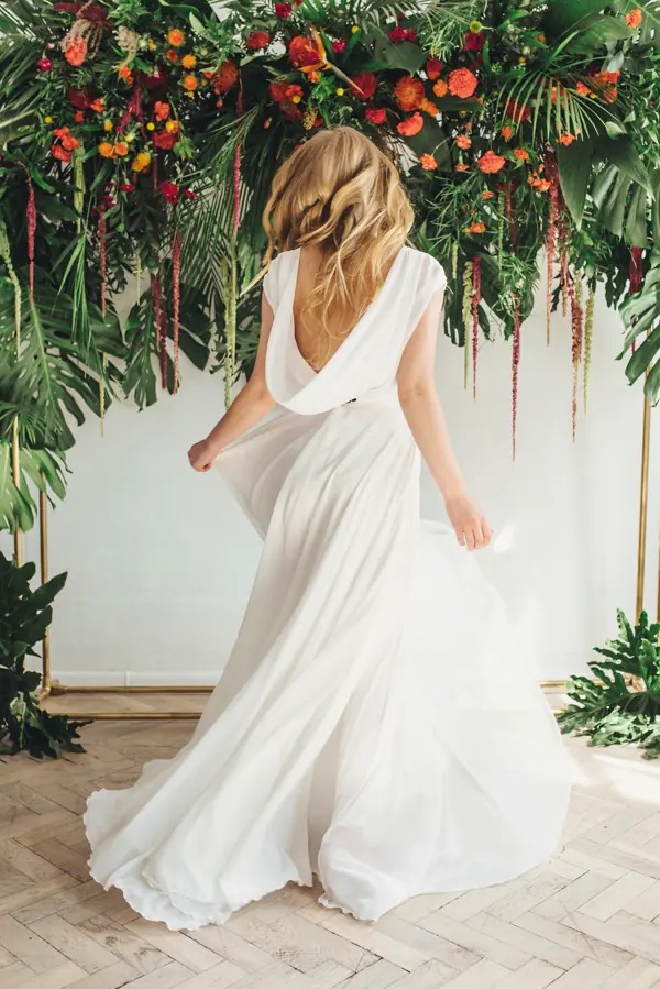 Kobieta w białej sukni ślubnej z odkrytymi plecami, tyłem do widza, na tle dekoracji z tropikalnych liści i kwiatów. Suknia powiewa, podłoga z jasnych desek.