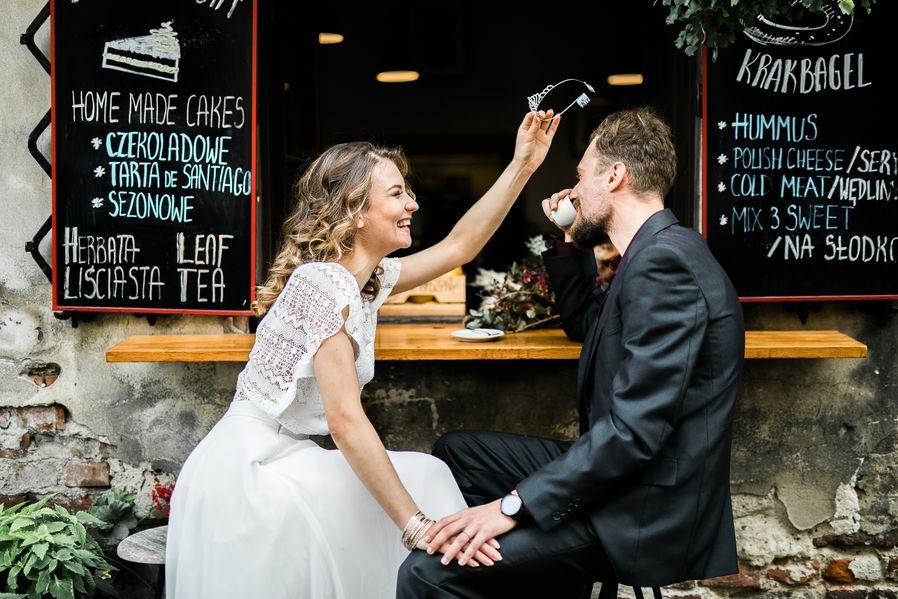 Panna młoda w białej sukni i koronkowym topie przymierza diadem mężczyźnie pijącemu kawę na tle menu kawiarni Krakbagel.