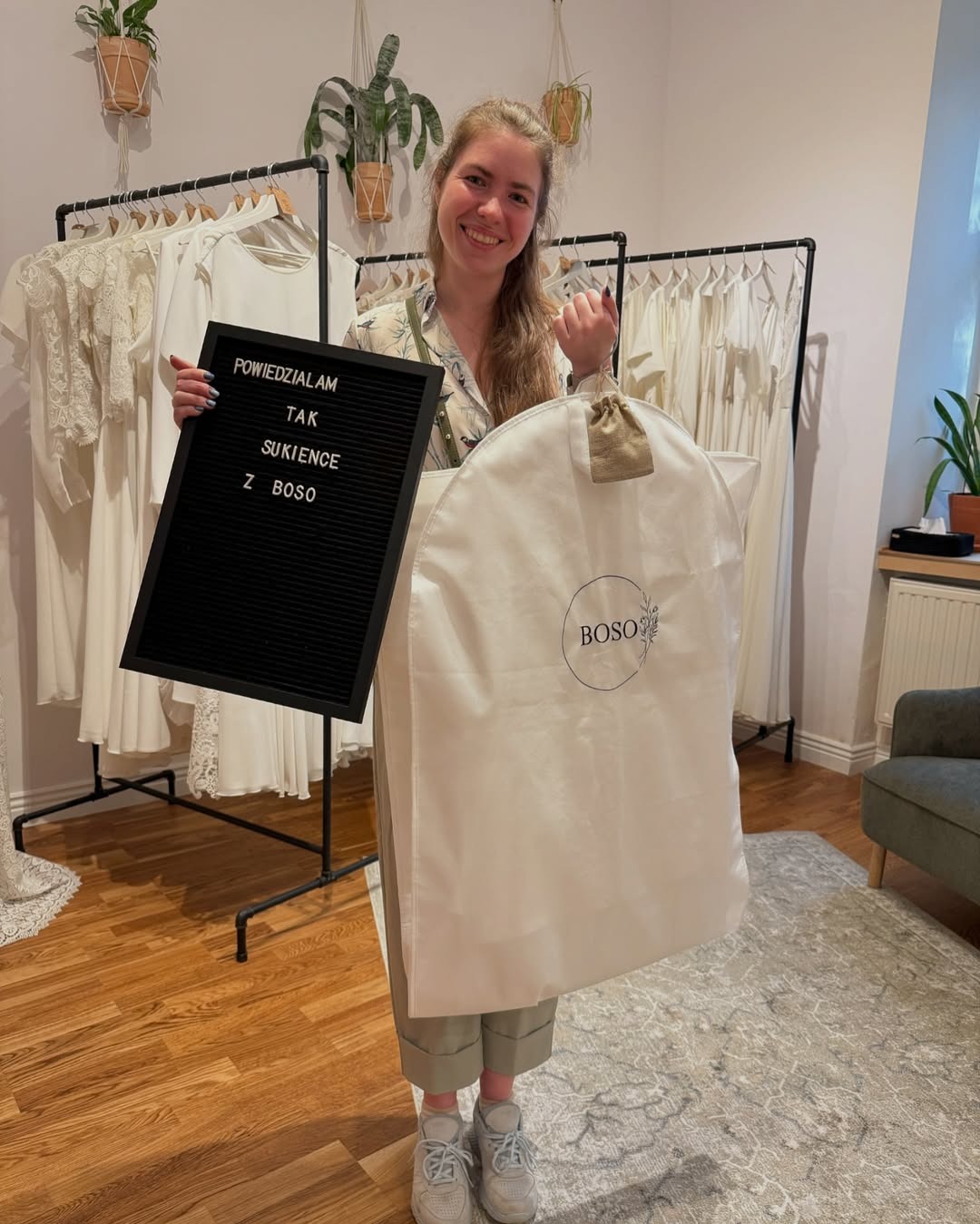 Uśmiechnięta kobieta trzyma torbę z logo BOSO i tabliczkę 'Powiedziałam TAK sukience Z BOSO' na tle wieszaków z sukniami ślubnymi.