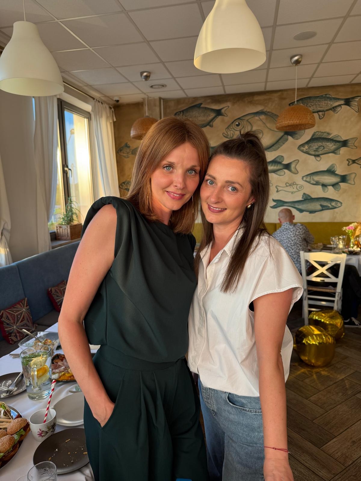 Dwie uśmiechnięte kobiety w eleganckich strojach, w restauracyjnym wnętrzu z motywem ryb na ścianie. Kobieta po lewej w ciemnozielonym kombinezonie, po prawej w białej koszuli i jeansach.