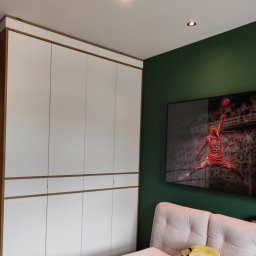 ROYAL MEBLE - Biała szafa wnękowa z drewnianymi akcentami na tle zielonej ściany z telewizorem, na którym wyświetlany jest obraz Michaela Jordana.
