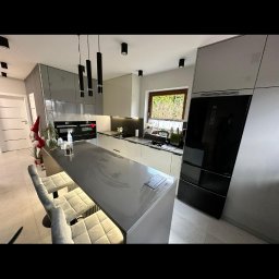 ROYAL MEBLE - Nowoczesna kuchnia z wyspą i hokerami w szaro-białej kolorystyce. Czarne lampy wiszące nad blatem. Minimalistyczny design z elementami dekoracyjnymi.