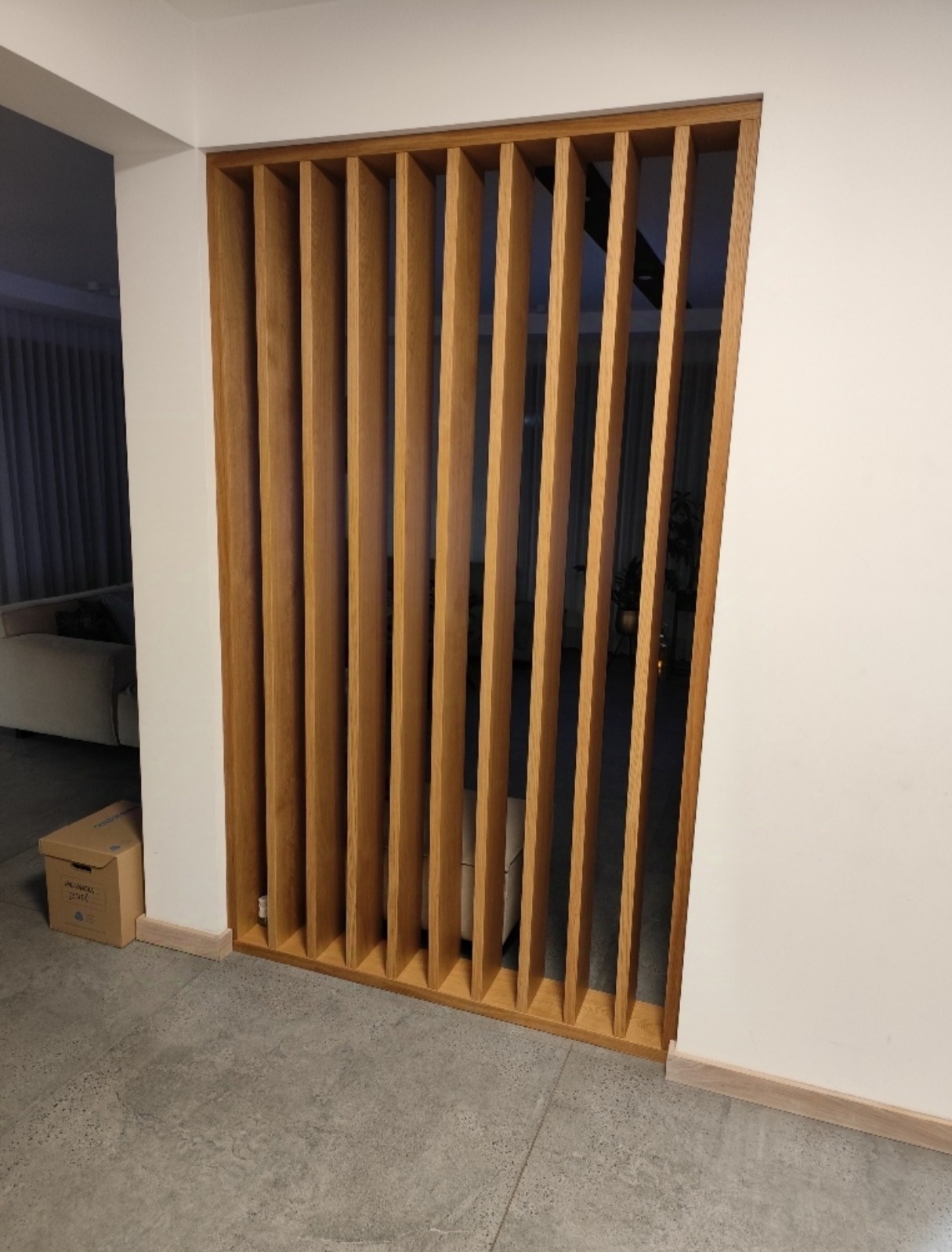 Drewniana ścianka działowa z pionowych listew, oddzielająca pomieszczenia. Widoczne szare płytki podłogowe i fragment sofy w tle. Minimalistyczny design.