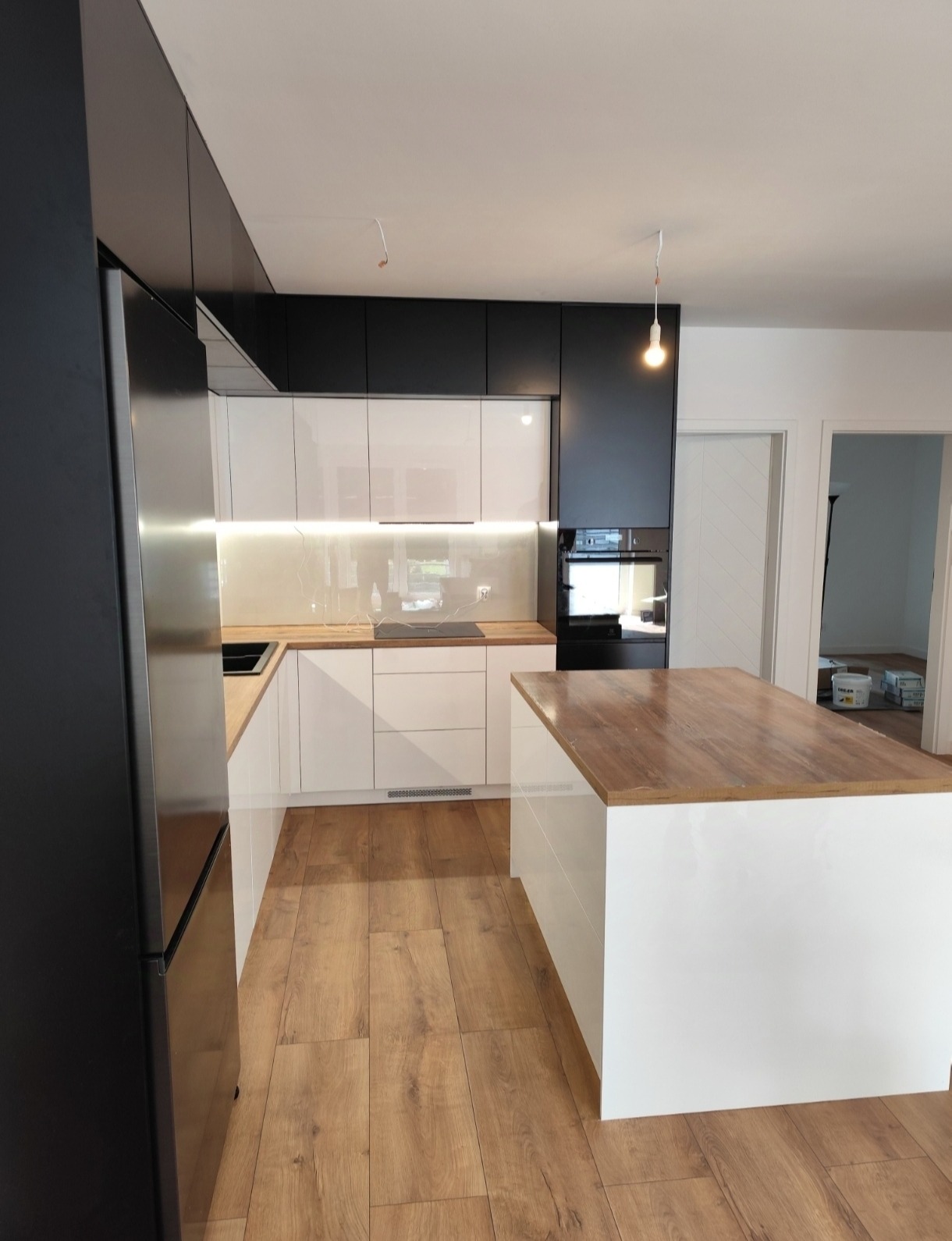 Nowoczesna kuchnia: białe szafki z drewnianym blatem i wyspą, czarne górne szafki, oświetlenie LED pod szafkami, drewniana podłoga, widok na otwarte drzwi.