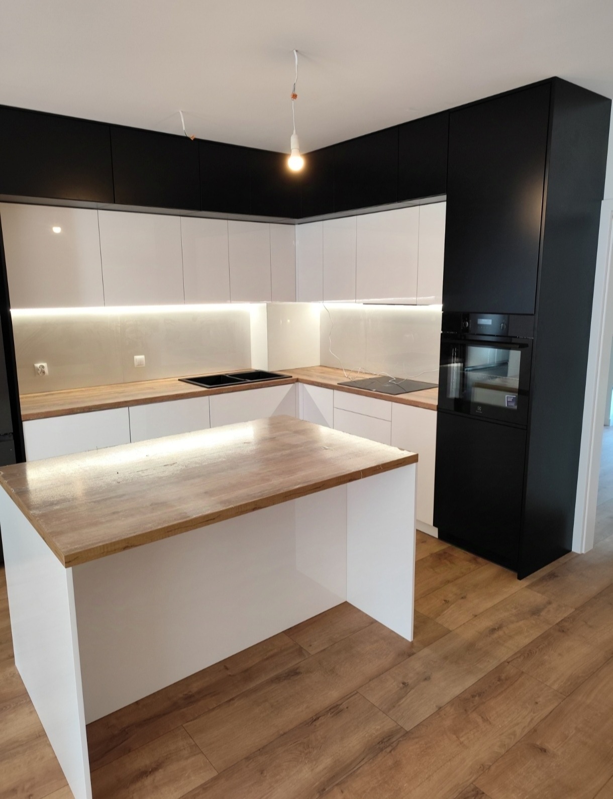 Nowoczesna kuchnia z wyspą: białe szafki dolne, czarne górne, drewniany blat wyspy, płyta indukcyjna i piekarnik w zabudowie. Oświetlenie LED pod szafkami.