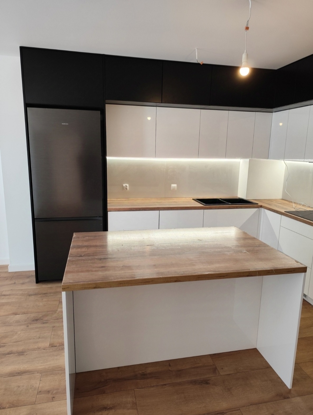 Nowoczesna kuchnia z wyspą: białe szafki, czarne górne szafki, drewniany blat wyspy i duża lodówka Samsung. Oświetlenie LED pod szafkami.
