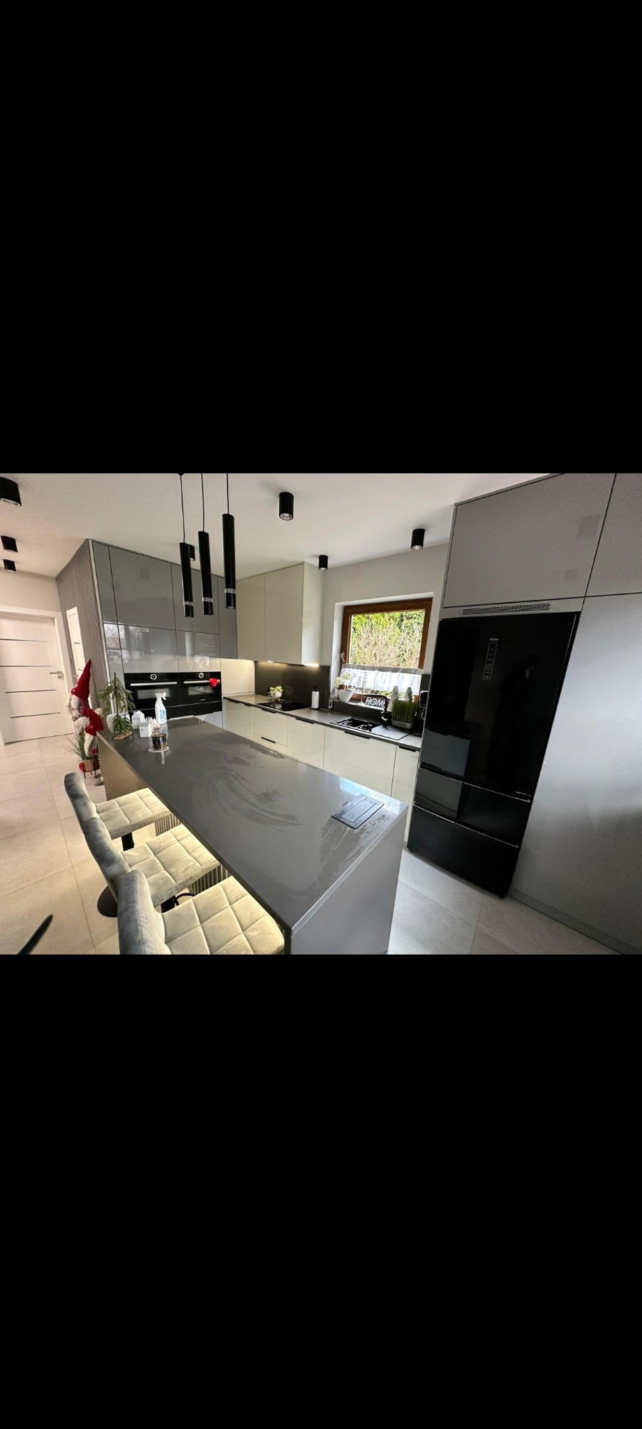 Nowoczesna kuchnia z wyspą i hokerami w szaro-białej kolorystyce. Czarne lampy wiszące nad blatem. Minimalistyczny design z elementami dekoracyjnymi.