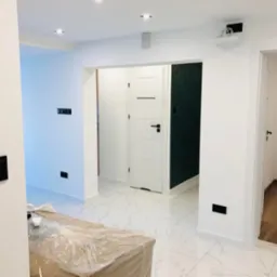 Białe, wykończone wnętrze z marmurową podłogą, czarnymi włącznikami i drzwiami. Częściowo zakryte folią meble sugerują świeżo zakończony remont.