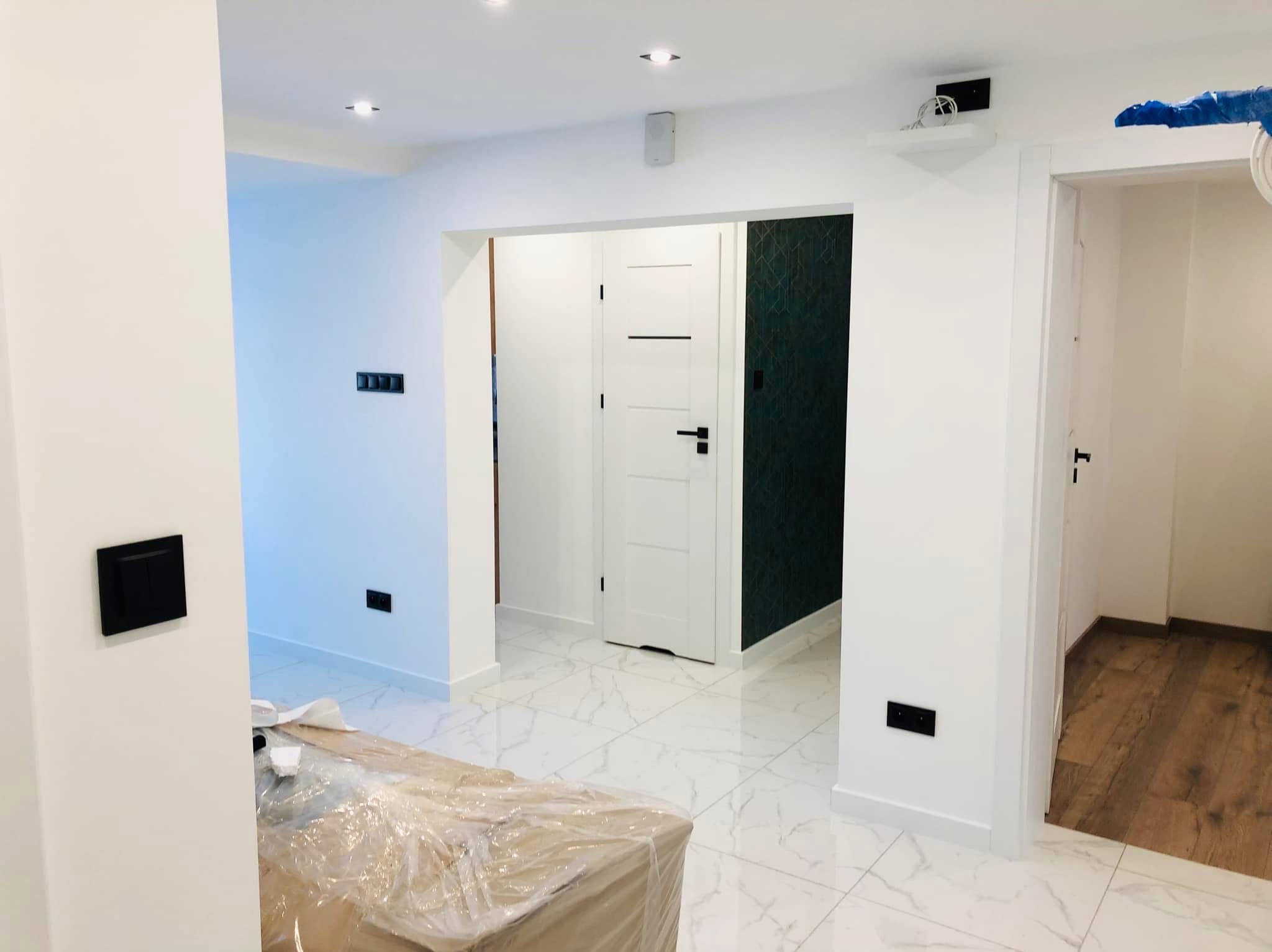 Białe, wykończone wnętrze z marmurową podłogą, czarnymi włącznikami i drzwiami. Częściowo zakryte folią meble sugerują świeżo zakończony remont.