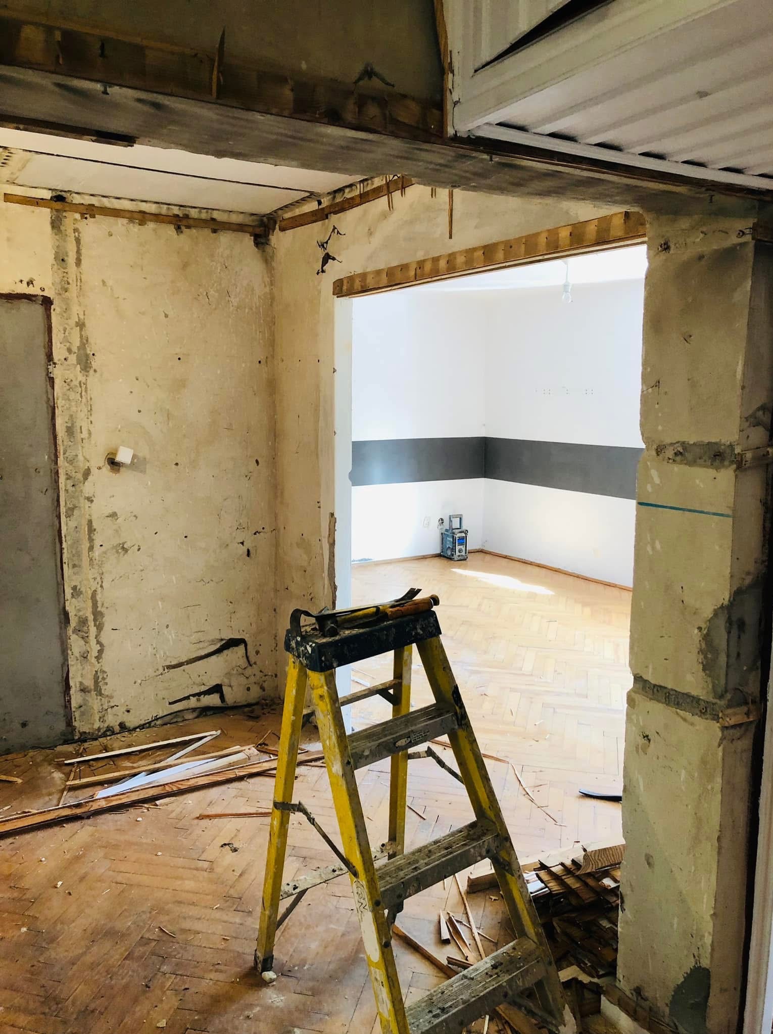 Wnętrze w trakcie demontażu: żółta drabina oparta o filar, odsłonięte fragmenty ścian i sufitu, parkiet z desek. W tle widoczne odnowione pomieszczenie.
